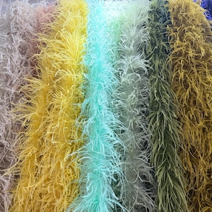 May include: A collection of long, fluffy feather boas arranged side by side in a vertical display. The colours include light blue, beige, bright yellow, mint green, pale green, olive green, mustard yellow, and deep navy blue, creating a vibrant gradient.