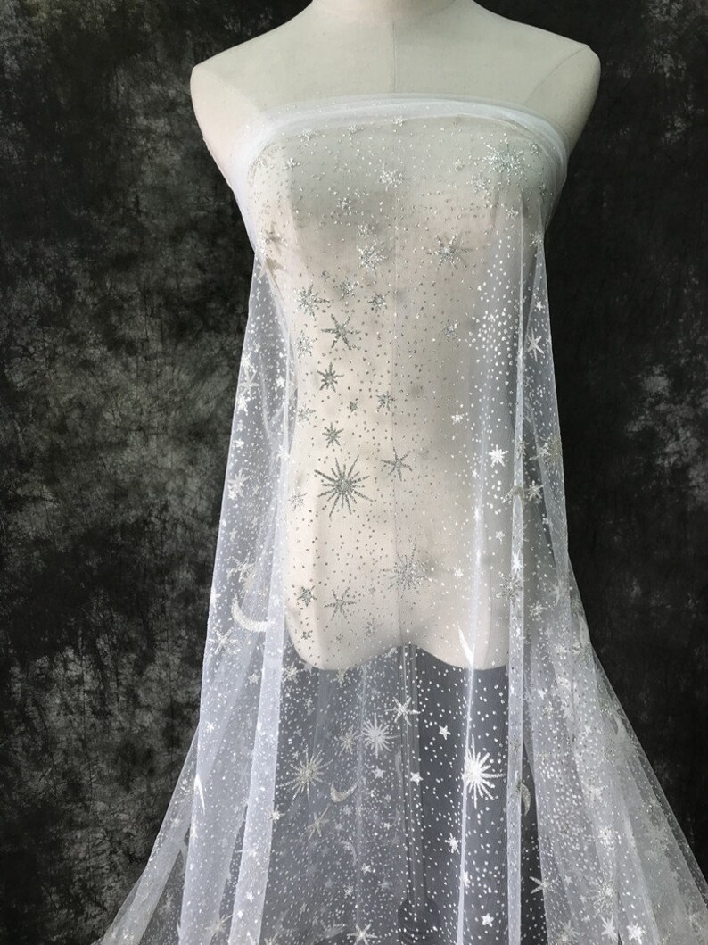 May include: White sheer fabric with a silver star and moon pattern. The fabric is draped over a mannequin, showcasing the delicate, shimmering design. The stars vary in size and are evenly distributed across the material.