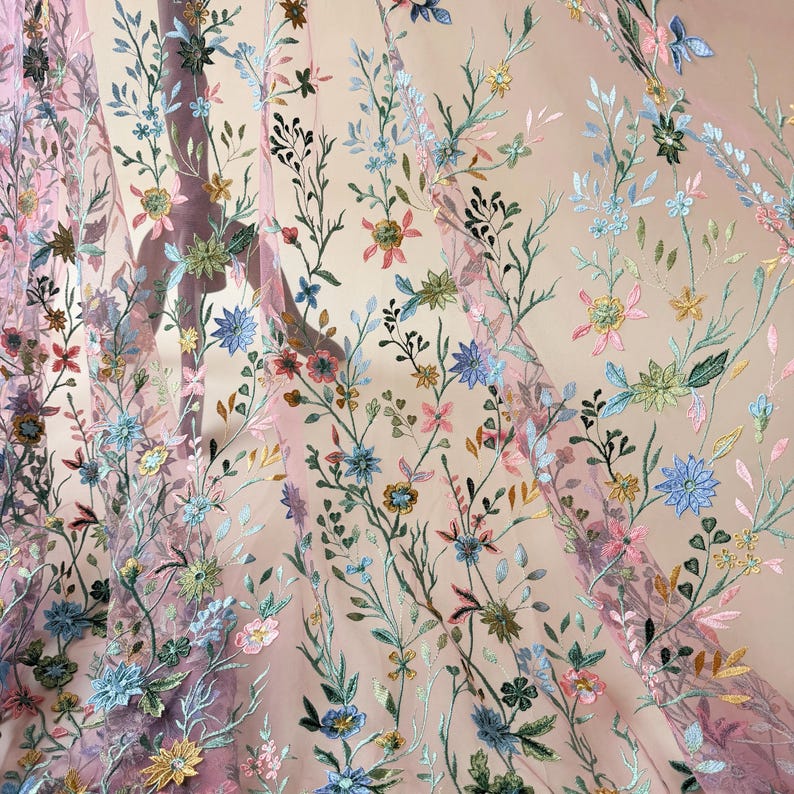 May include: Sheer pink fabric with embroidered floral patterns. The embroidery features a variety of colorful flowers in shades of blue, pink, yellow, and green, with delicate green leaves and stems. The fabric is semi-transparent.