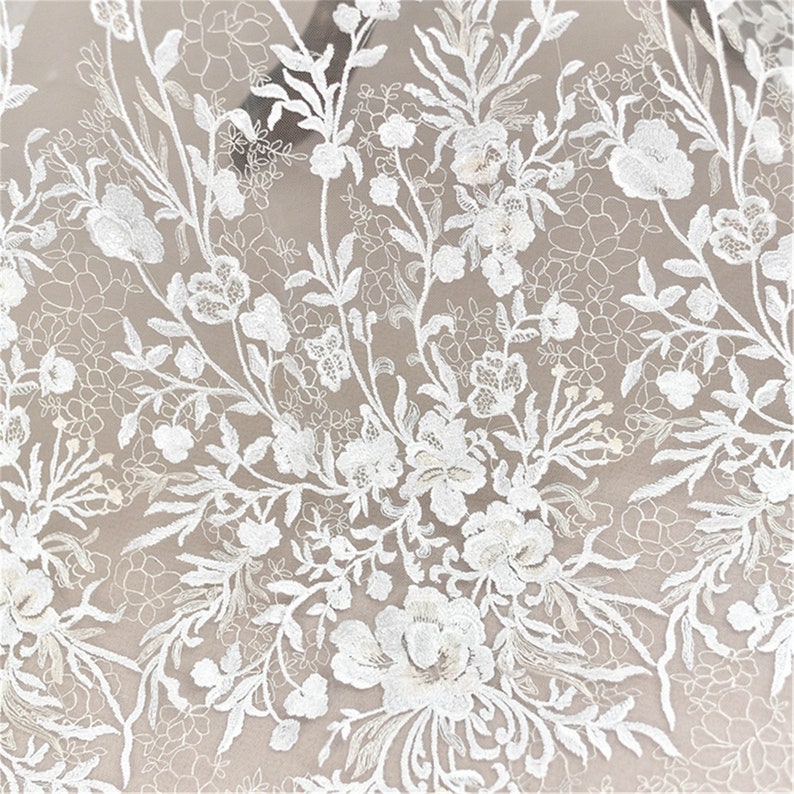 May include: White floral lace fabric with a delicate pattern of flowers and leaves. The lace is sheer and has a soft, romantic feel.
