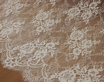 May include: Ivory lace fabric with a floral pattern. The delicate lace features detailed floral designs and a scalloped edge. The fabric is semi-transparent, allowing the background to be partially visible. Ideal for bridal wear, dresses, or decorative projects.