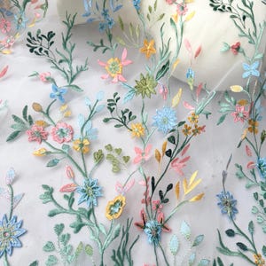 May include: Delicate floral embroidery on a white sheer fabric. The design features colorful embroidered flowers in shades of pink, yellow, blue, and green, with detailed leaves and stems. The embroidery creates a textured, three-dimensional effect, perfect for apparel or home decor.