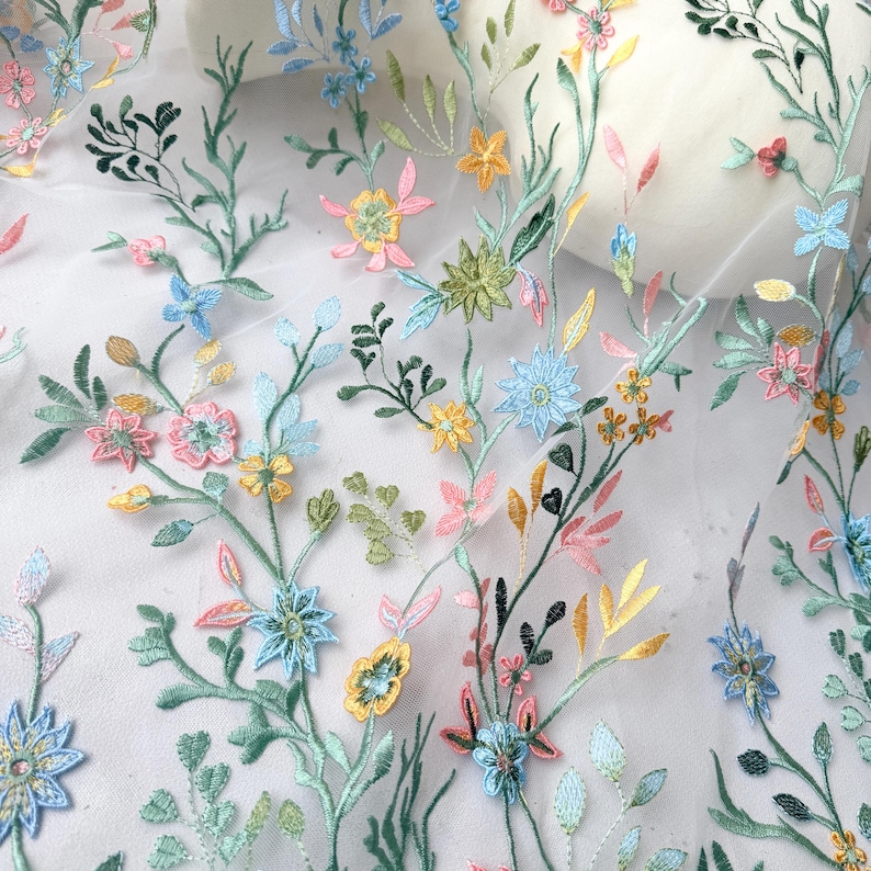 May include: Delicate floral embroidery on a white sheer fabric. The design features colorful embroidered flowers in shades of pink, yellow, blue, and green, with detailed leaves and stems. The embroidery creates a textured, three-dimensional effect, perfect for apparel or home decor.