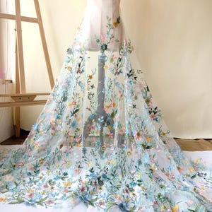 May include: Sheer white fabric with embroidered floral design. The embroidery features a variety of colorful flowers in shades of blue, pink, yellow, and green, with delicate green stems and leaves. The fabric is draped over a mannequin.