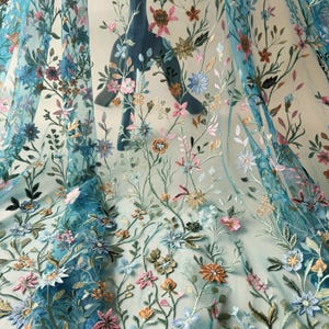 May include: A sheer turquoise fabric with intricate floral embroidery. The design features a variety of colorful flowers in shades of pink, blue, orange, and gold, with green leaves and stems. The fabric is likely used for apparel or decorative purposes.