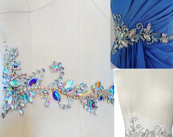 May include: A close-up of a beaded embellishment featuring iridescent crystals arranged in a floral pattern. The embellishment is shown on three different surfaces, including a white mannequin torso, a blue fabric background, and a white fabric background.