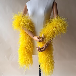 May include: A vibrant yellow feather boa draped over a white mannequin torso. The long, fluffy accessory features dense, bright yellow plumage that creates a voluminous texture, extending from the shoulders down to the waist area of the form.