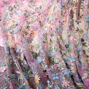 May include: Close-up of a sheer, floral embroidered fabric. The fabric features a delicate pattern of colorful flowers and green leaves on a pink and purple background. The embroidery includes blue, yellow, and pink flowers.