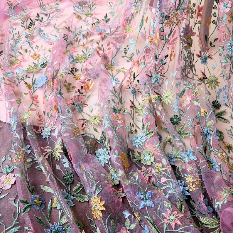 May include: Close-up of a sheer, floral embroidered fabric. The fabric features a delicate pattern of colorful flowers and green leaves on a pink and purple background. The embroidery includes blue, yellow, and pink flowers.