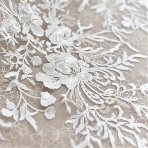 May include: White floral lace fabric with sequins. The lace features a delicate floral pattern with intricate details. The fabric is sheer and lightweight, making it ideal for a variety of projects.