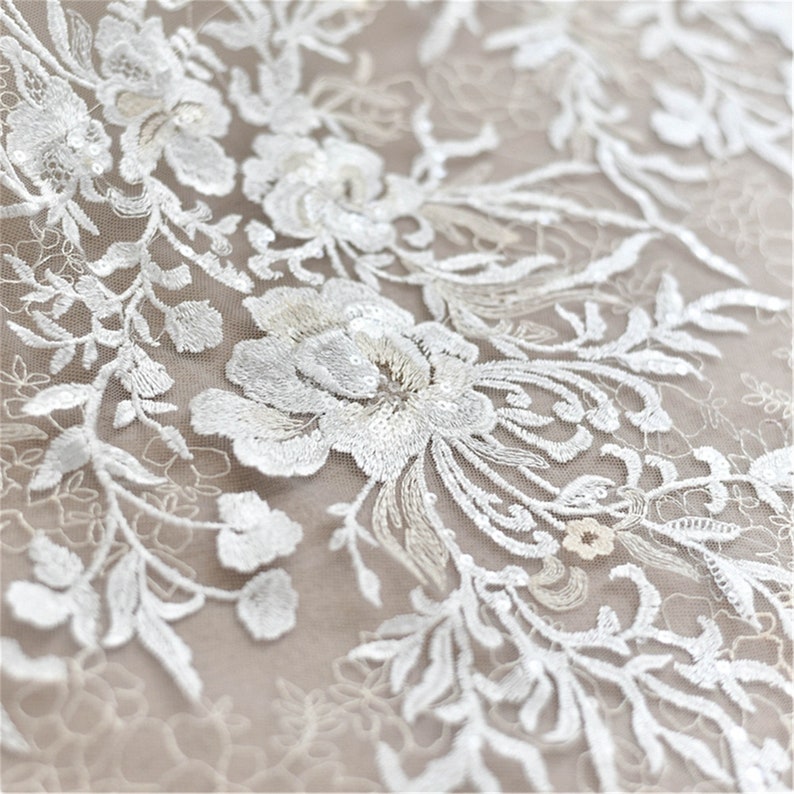May include: White floral lace fabric with sequins. The lace features a delicate floral pattern with intricate details. The fabric is sheer and lightweight, making it ideal for a variety of projects.