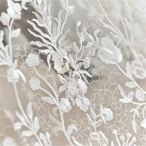 May include: White floral lace fabric with a delicate pattern of flowers and leaves. The lace is sheer and has a soft, romantic feel.
