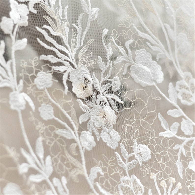 May include: White floral lace fabric with a delicate pattern of flowers and leaves. The lace is sheer and has a soft, romantic feel.