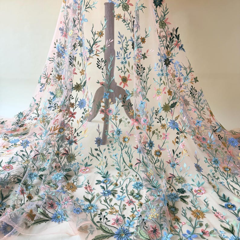 May include: Delicate floral embroidered fabric in shades of pink, blue, green, and gold. The embroidery features a variety of flowers and leaves, creating a textured and dimensional effect. The fabric is sheer, allowing light to pass through.