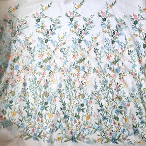 May include: White fabric with embroidered floral design. The embroidery features a variety of colorful flowers in shades of yellow, pink, blue, and green, with delicate green stems and leaves. The fabric is likely used for apparel or home decor.