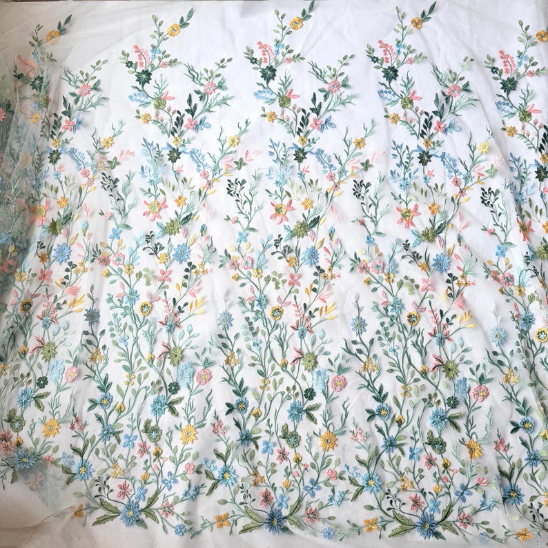 May include: White fabric with embroidered floral design. The embroidery features a variety of colorful flowers in shades of yellow, pink, blue, and green, with delicate green stems and leaves. The fabric is likely used for apparel or home decor.