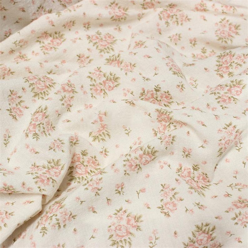 May include: Off-white fabric with a repeating pattern of pink roses and green leaves. The floral design is scattered across the material, creating a delicate and romantic aesthetic. The fabric appears soft and slightly textured.