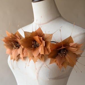 Könnte beinhalten: Ein dekoratives Accessoire bestehend aus drei großen, geschichteten Blumen aus hellbraunen Federn. Jede Blume hat in der Mitte eine Ansammlung kleiner, metallisch goldener Perlen. Das Stück wird auf einem cremefarbenen Schneiderbüsten-Torso präsentiert.