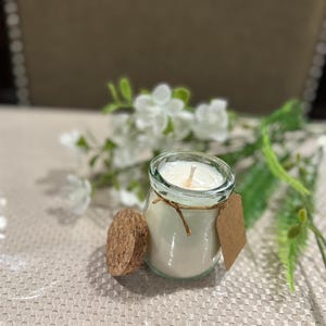 May include: A small glass jar candle with white wax and a single wick. The jar has a cork lid and a brown paper tag tied around the top.