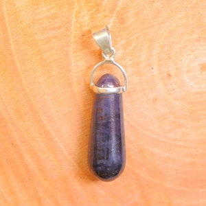 Puede incluir: Un colgante de plata con una piedra preciosa de color púrpura oscuro en forma de lágrima. El colgante está colgado de una cadena de plata.