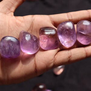 May include: Six smooth, polished amethyst stones in various shades of purple, ranging from light lavender to deep violet, are displayed in a hand.