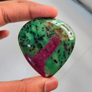 May include: A green and pink gemstone with black flecks. The gemstone is heart-shaped and has a smooth, polished surface.