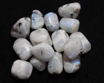 Op de afbeelding: Een verzameling witte maansteen-tumbled stenen met iriserende blauwe en zwarte vlekken. De stenen zijn glad en gepolijst en liggen op een zwarte achtergrond.