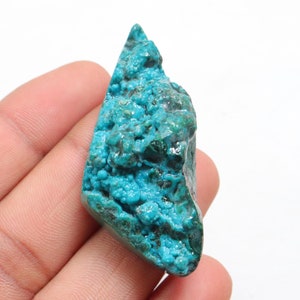 May include: A rough, teal blue, natural gemstone with a crystalline texture. The stone is irregularly shaped and has a pointed top.
