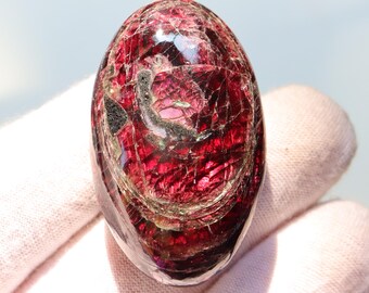 Könnte beinhalten: Ein polierter, oval geformter Edelstein in tiefroter Farbe, der innere Muster und Einschlüsse aufweist. Die Oberfläche des Steins reflektiert das Licht und hebt seine kristalline Struktur hervor. Der Edelstein wird in einer behandschuhten Hand gehalten.