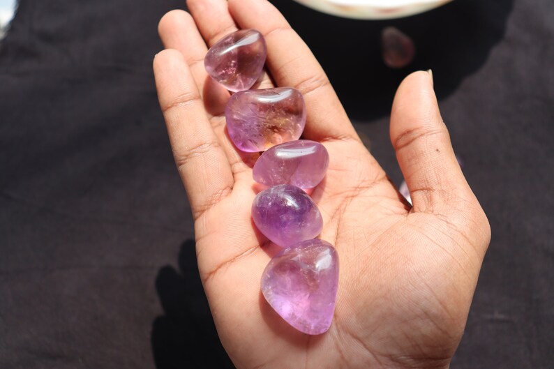 May include: Five smooth, polished amethyst stones in various shades of purple, arranged in a row on a hand.