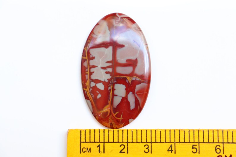 May include: A red, white, and gold oval-shaped cabochon gemstone with a unique, abstract pattern. The gemstone is approximately 3 cm long and is displayed next to a ruler.