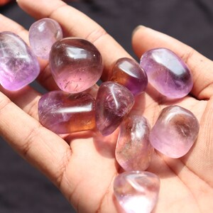 May include: A collection of tumbled amethyst stones in various shades of purple and lavender. The stones are smooth and polished, with a natural, organic shape.