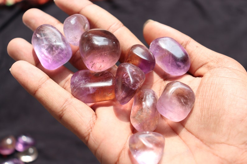 May include: A collection of tumbled amethyst stones in various shades of purple and lavender. The stones are smooth and polished, with a natural, organic shape.
