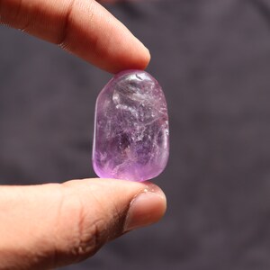 May include: A smooth, polished amethyst gemstone with a light purple hue. The gemstone is oval-shaped and has a translucent appearance.