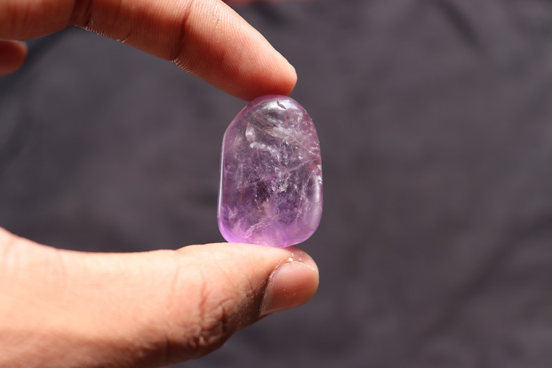 May include: A smooth, polished amethyst gemstone with a light purple hue. The gemstone is oval-shaped and has a translucent appearance.
