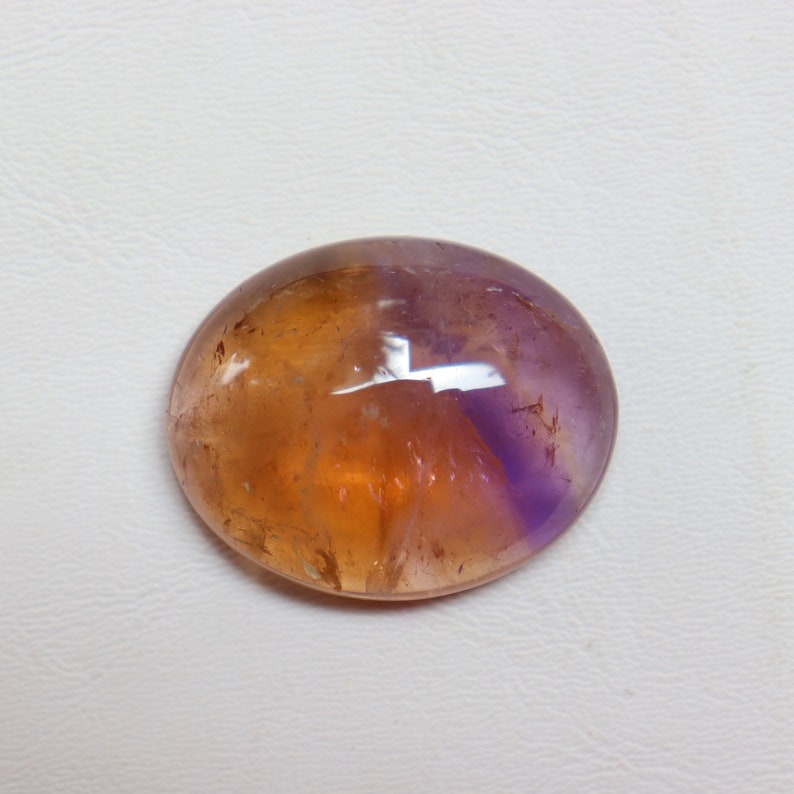 May include: A smooth, oval-shaped gemstone with a bi-color pattern. The top half is a light purple, while the bottom half is a warm amber color.