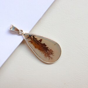 May include: A silver pendant featuring a teardrop-shaped dendritic agate stone with brown and white patterns. The stone is set in a silver bezel.