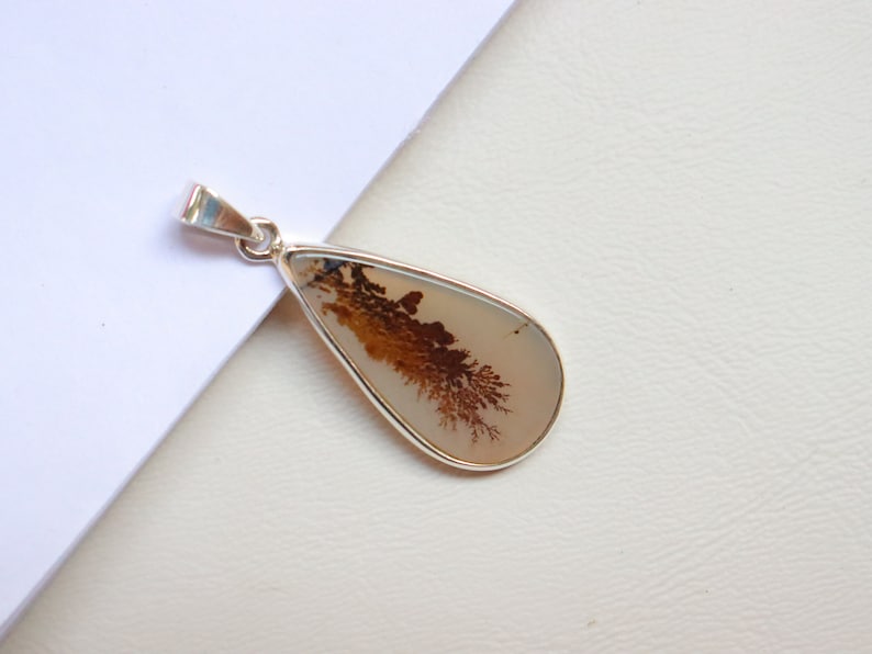 May include: A silver pendant featuring a teardrop-shaped dendritic agate stone with brown and white patterns. The stone is set in a silver bezel.