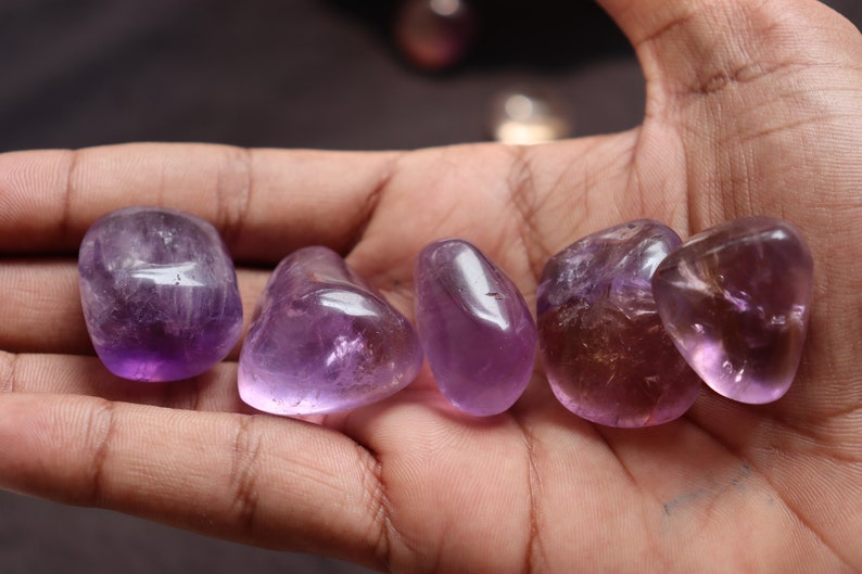 May include: Five smooth, polished amethyst stones in various shades of purple, ranging from light lavender to deep violet. The stones are tumbled and have a natural, organic shape.