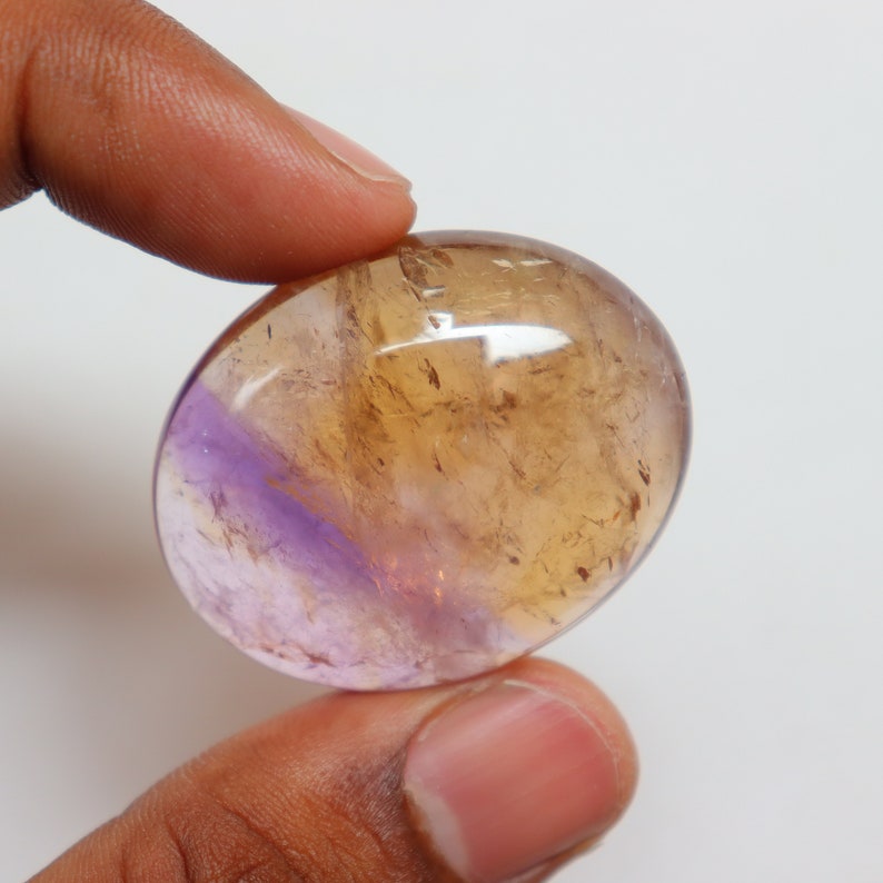 May include: A smooth, oval-shaped gemstone with a layered appearance. The top half is a translucent golden yellow, while the bottom half is a translucent purple.