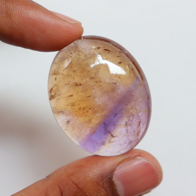 May include: A smooth, oval-shaped gemstone with a light brown and purple color. The gemstone has a translucent appearance with a slight shimmer.