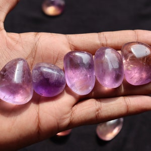 May include: Six smooth, polished amethyst stones in various shades of purple, arranged in a row on a hand.