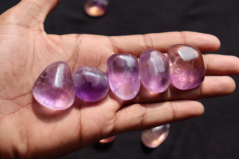 May include: Six smooth, polished amethyst stones in various shades of purple, arranged in a row on a hand.