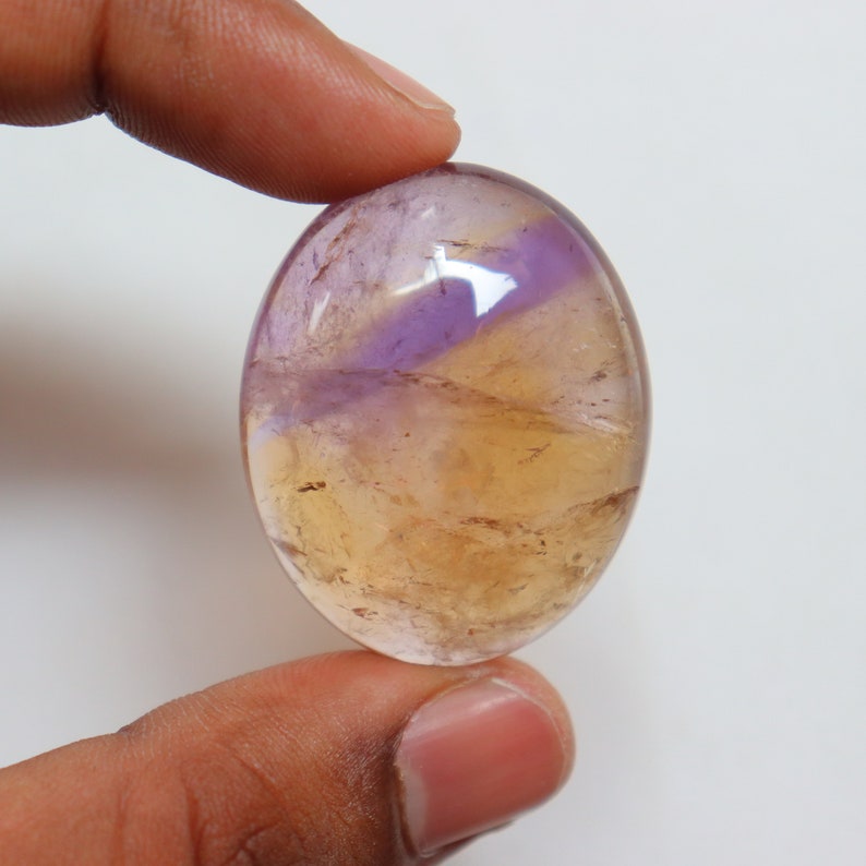 May include: A smooth, oval-shaped, cabochon gemstone with a yellow and purple color pattern. The stone is translucent and has a glossy finish.