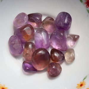May include: A collection of tumbled amethyst and citrine stones in various shades of purple and orange. The stones are smooth and polished, and they are arranged in a bowl.