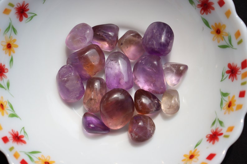 May include: A collection of tumbled amethyst and citrine stones in various shades of purple and orange. The stones are smooth and polished, and they are arranged in a bowl.