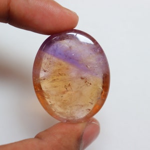 May include: A smooth, oval-shaped gemstone with a purple and yellow color gradient. The stone is translucent and has a slight shimmer.