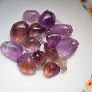 May include: A collection of tumbled amethyst and amethyst citrine stones in various shades of purple and yellow. The stones are smooth and polished, and they are arranged in a small white bowl with a floral pattern.