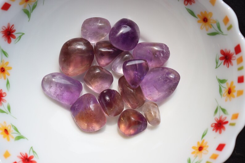 May include: A collection of tumbled amethyst and amethyst citrine stones in various shades of purple and yellow. The stones are smooth and polished, and they are arranged in a small white bowl with a floral pattern.