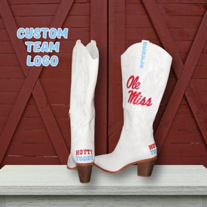 May include: A pair of white leather cowboy boots featuring the red text Ole Miss and the blue text Hotty Toddy. The boots have a stacked brown heel and subtle tonal stitching patterns. The text Custom Team Logo is visible in the upper left corner.
